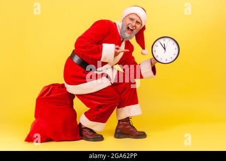 Staunender älterer Mann mit grauem Bart im weihnachtsmannkostüm sitzt auf einer großen roten Tasche mit Geschenken zu Weihnachten und zeigt voller Aufregung auf die Wanduhr. Stockfoto