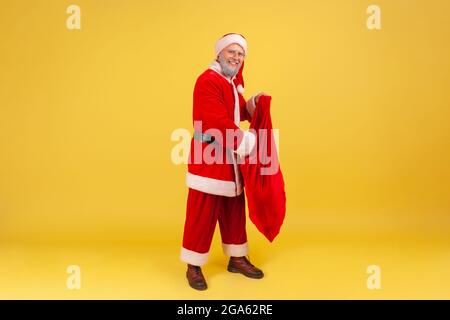 Lächelnder älterer Mann mit grauem Bart trägt weihnachtsmannkostüm und steckt die Hand in eine große rote Tasche voller Geschenke für Weihnachten, Gruß mit Neujahr Stockfoto