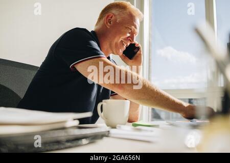 Älterer Mann, der am Schreibtisch im Büro sitzt und mit einem Smartphone telefoniert. Leitender Geschäftsmann, der im Büro arbeitet und mit dem Mobiltelefon spricht. Stockfoto