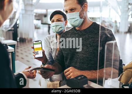 Touristenfamilie, die während einer Pandemie am Check-in-Schalter am Flughafen den Impfpass vorzeigt. Mann, der am Check-in-Schalter steht, während Frau digitale V-Anzeigen vorführt Stockfoto