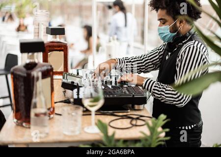 Afrikanischer dj, der in der Cocktailbar im Freien Musik spielt, während er eine Gesichtsschutzmaske trägt - Hauptaugenmerk liegt auf der Oberhand Stockfoto