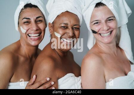 Reife Frauen lachen zusammen, während sie Schönheitsbehandlungen im Spa mit Hautmaske machen - Hauptfokus auf afrikanisches weibliches Gesicht - Hauptfokus auf rechtes Hausgesicht Stockfoto