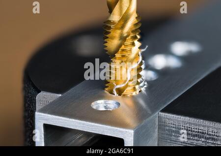 Spiralförmiger, geriffelter Stahlzapfer, der ein Gewinde in der Bohrung des Alu-Profils macht, das in den Schraubbacken eingespannt ist. Titan beschichtetes goldenes Schneidwerkzeug und verdrehte Späne Detail. Stockfoto