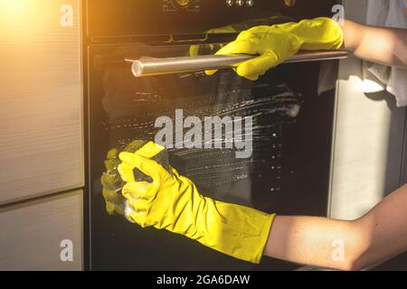 Elektroofen in moderner Küche waschen, gelbe Gummihandschuhe und Schwamm in der Hand Stockfoto
