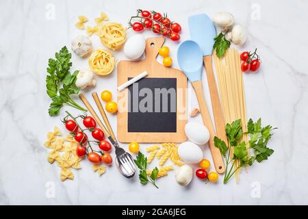 Kleine Tafel mit Kopierplatz umgeben von Zutaten zum Kochen verschiedene Arten von Pasta, Gewürze, Utensilien, gesunde rohe Gemüse Scattere Stockfoto