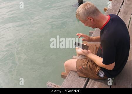 Ein erwachsener Mann aus dem Kaukasus, der sein Smartphone benutzt, während er auf einem Pier sitzt. Stockfoto