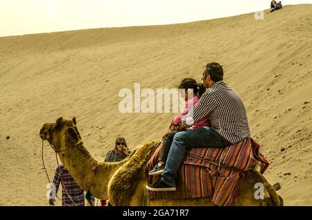 Yazd, Wüste, Iran, 20. Februar 2021: Mann mit einem kleinen Mädchen, das auf einem mit einem hellen Teppich bedeckten Kamel sitzt, begleitet von einem Führer und einer Wattin Stockfoto