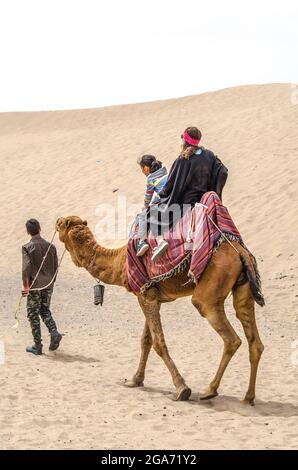 Yazd, Wüste, Iran, 20. Februar 2021: Eine junge Frau und ein kleines Mädchen sitzen auf einem Kamel, bedeckt mit einem Teppich, begleitet von einem Führer, bewegen sich durch die Stockfoto