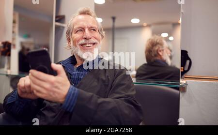 Älterer Mann Wartet Auf Haarschnitt Im Friseursalon Mit Blick Auf Mobiltelefon Stockfoto