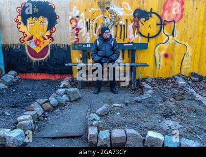 Älterer Mann, der auf einer Bank neben einer farbenfrohen Wand im Garten der informellen Hausbesetzer-Gemeinde in Kreuzberg-Berlin sitzt. Stockfoto