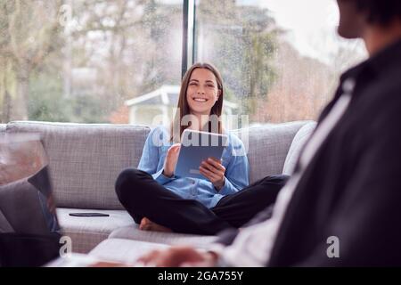 Entspanntes Junges Paar Zu Hause Auf Dem Sofa Sitzend Mit Digitalem Tablet Und Lesebuch Stockfoto