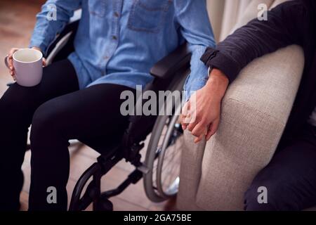 Nahaufnahme Eines Paares Zu Hause Mit Einer Behinderten Frau Im Rollstuhl, Die Hände Hält, Während Der Mann Auf Dem Sofa Sitzt Stockfoto