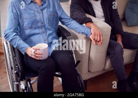Nahaufnahme Eines Paares Zu Hause Mit Einer Behinderten Frau Im Rollstuhl, Die Hände Hält, Während Der Mann Auf Dem Sofa Sitzt Stockfoto
