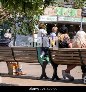 Epsom Surrey London, Juli 29 2021, Gruppe der Anonymen Männer und Frauen, die auf der Bank sitzen und an den Käufern vorbeilaufen Stockfoto