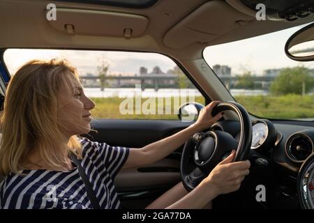 Porträt einer unbeschwerten europäischen mittleren Frau, die im Auto fährt, lächelnd, glücklich während der Fahrt bei Sonnenuntergang Stockfoto