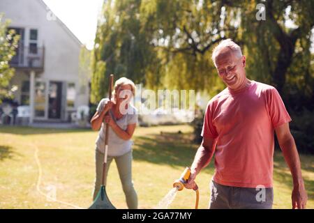 Rentnerpaar Bei Der Arbeit Bewässerung Von Pflanzen Mit Schlauch Und Aufräumen Garten Mit Rake Zu Hause Stockfoto