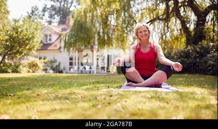 Porträt Einer Älteren Frau Zu Hause Im Garten, Die Fitnesskleidung Trägt, Die Auf Einer Yogamatte Sitzt Stockfoto