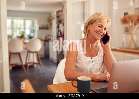 Pensionierte Frau Am Telefon Zu Hause In Der Küche Mit Laptop Stockfoto