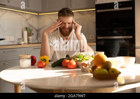 Junger, müder Mann mit Kopfschmerzen, der am Tisch sitzt Stockfoto