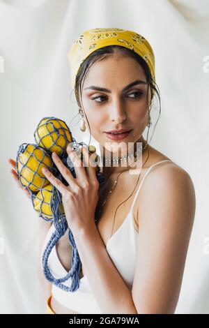 Frau in gelbem Kopftuch und Crop Top hält String-Tasche mit Zitronen auf weiß Stockfoto