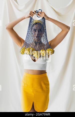 Frau in Crop Top hält Saitentasche mit Zitronen auf weiß Stockfoto