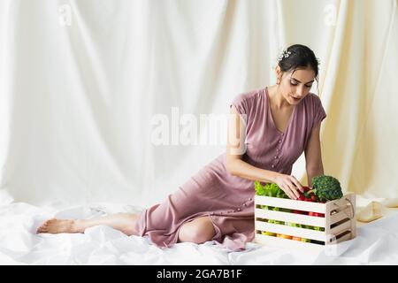 Brunette Frau erreicht roten Paprika in der Nähe von Gemüse in Holzbehälter auf weiß Stockfoto