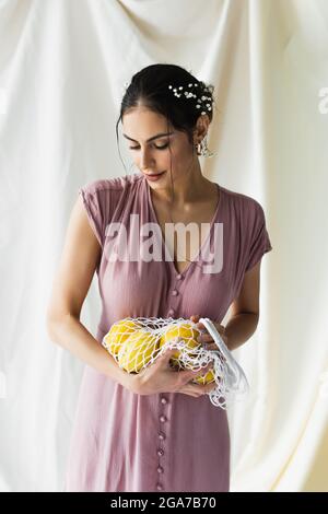 Brünette Frau mit Blumen im Haar hält wiederverwendbare Saitenbeutel mit Zitronen auf weiß Stockfoto