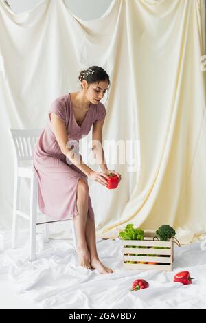 Brunette Frau hält roten Paprika in der Nähe von Gemüse in Holzbehälter auf weiß Stockfoto