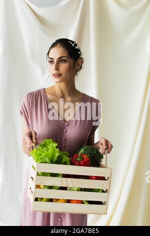 Brunette Frau hält Holzkiste mit Gemüse auf weiß Stockfoto