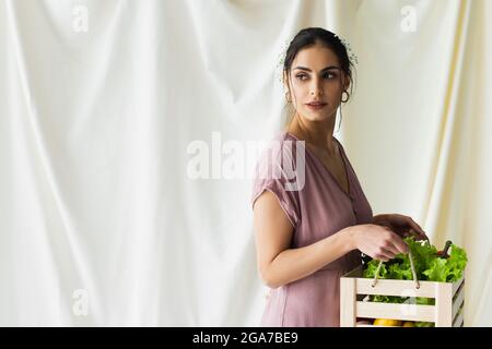 Hübsche Frau, die eine Holzkiste mit Gemüse auf Weiß hält Stockfoto