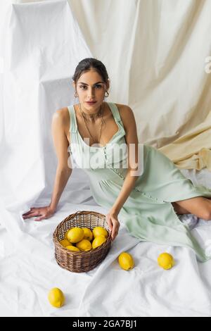 High-Angle-Ansicht der hübschen Frau im Kleid sitzt in der Nähe Korbkorb mit Zitronen auf weiß Stockfoto