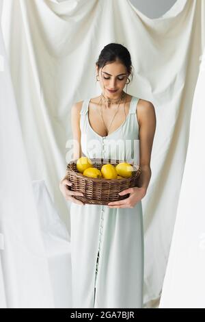Lächelnde Frau in Kleid suchen reifen Zitronen in Weidenkorb auf weiß Stockfoto