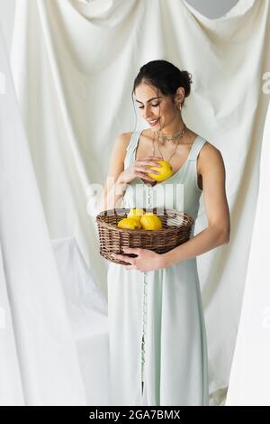 Erfreut Frau im Kleid hält reife Zitronen in Weidenkorb auf weiß Stockfoto