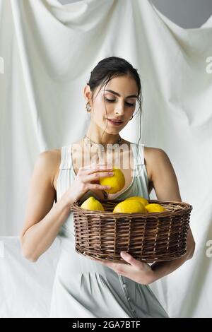 Lächelnde Frau in Kleid hält Korbkorb mit Zitronen auf weiß Stockfoto