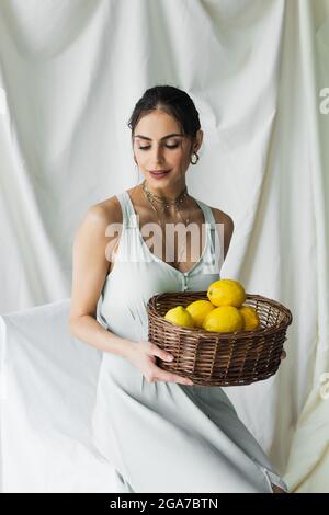 Glückliche Frau in Kleid hält Korbkorb mit Zitronen auf weiß Stockfoto