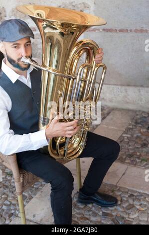 Ein Jazzmusiker im Vintage-Stil, der bei hellem Tageslicht auf der Straße Tuba spielt. Mit blau gefärbtem Bart und Ziegenbart und einer Retro-Mütze. Stockfoto