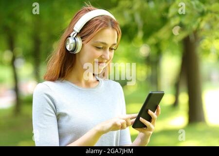 Eine zufriedene Frau, die Kopfhörer trägt und Musik auf einem Smartphone in einem Park hört Stockfoto