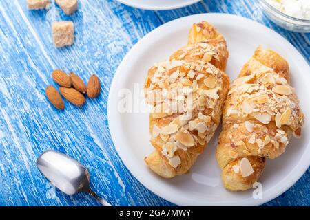 Frühstück am Morgen. Frisches französisches Croissant auf einem Teller. In der Nähe befinden sich verstreute Mandeln und Rohrzucker-Würfel auf einer blauen Holzoberfläche. Traditionelle französische Küche Stockfoto