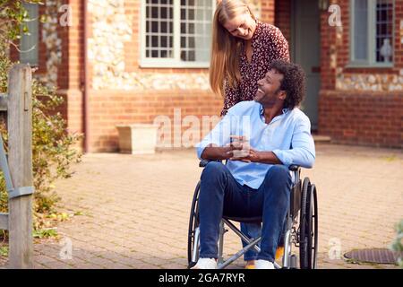 Reifes Paar Mit Mann, Der Im Rollstuhl Sitzt Und Von Einer Frau Außerhalb Des Hauses Geschoben Wird Stockfoto