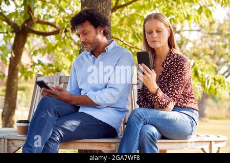 Reifes Paar Treffen Im Outdoor Park Auf Der Bank Sitzend Mit Blick Auf Mobiltelefone Stockfoto