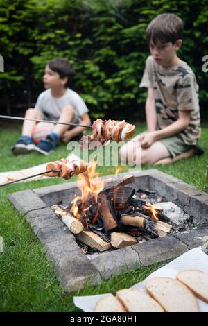 Zwei Scheiben Wurst, Speck und Zwiebel für Kinder, gebraten auf Lagerfeuer, mit verschwommenen Kindern im Hintergrund. Picknick, Grill, Kochkonzept. Sommer Stockfoto