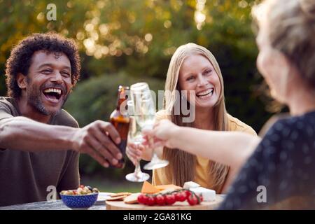 Eine Gruppe Von Freunden Feiert Mit Bier Und Champagner, Während Sie Am Tisch Im Garten Mit Snacks Sitzen Stockfoto