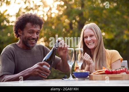 Reifer Mann Eröffnet Champagner-Flasche, Während Das Paar Mit Snacks Am Tisch Im Garten Sitzt Stockfoto