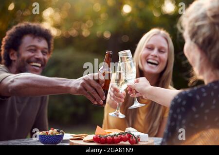Eine Gruppe Von Freunden Feiert Mit Bier Und Champagner, Während Sie Am Tisch Im Garten Mit Snacks Sitzen Stockfoto