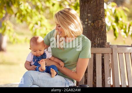 Liebende Mutter Kuscheln Und Spielen Mit Baby-Tochter Im Freien Sitzen Auf Sitz Unter Sommerbaum Stockfoto