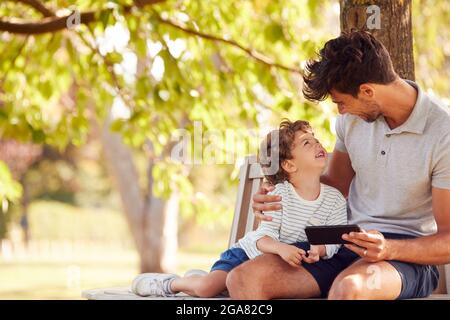 Vater Sitzt Auf Der Parkbank Unter Baum Mit Sohn Blick Auf Handy Zusammen Stockfoto