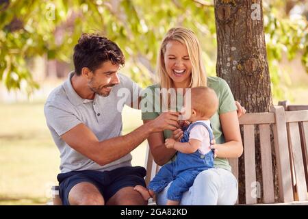 Familie Mit Baby-Tochter Sitzt Auf Seat Under Tree Im Sommer Park Zusammen Stockfoto