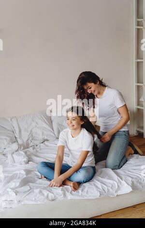 Mutter, die ihrer Tochter, die zu Hause auf dem Bett sitzt, Zöpfe herstellt. Stockfoto