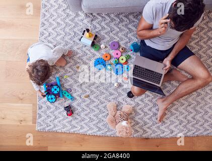 Overhead Shot Of Son Spielt Mit Spielzeug Auf Teppich Zu Hause, Während Vater Nutzt Laptop Und Handy Stockfoto