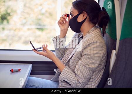 Geschäftsfrau im Zug beim Anlegen von Make-up während der Pandemie mit PSA-Gesichtsmaske Stockfoto